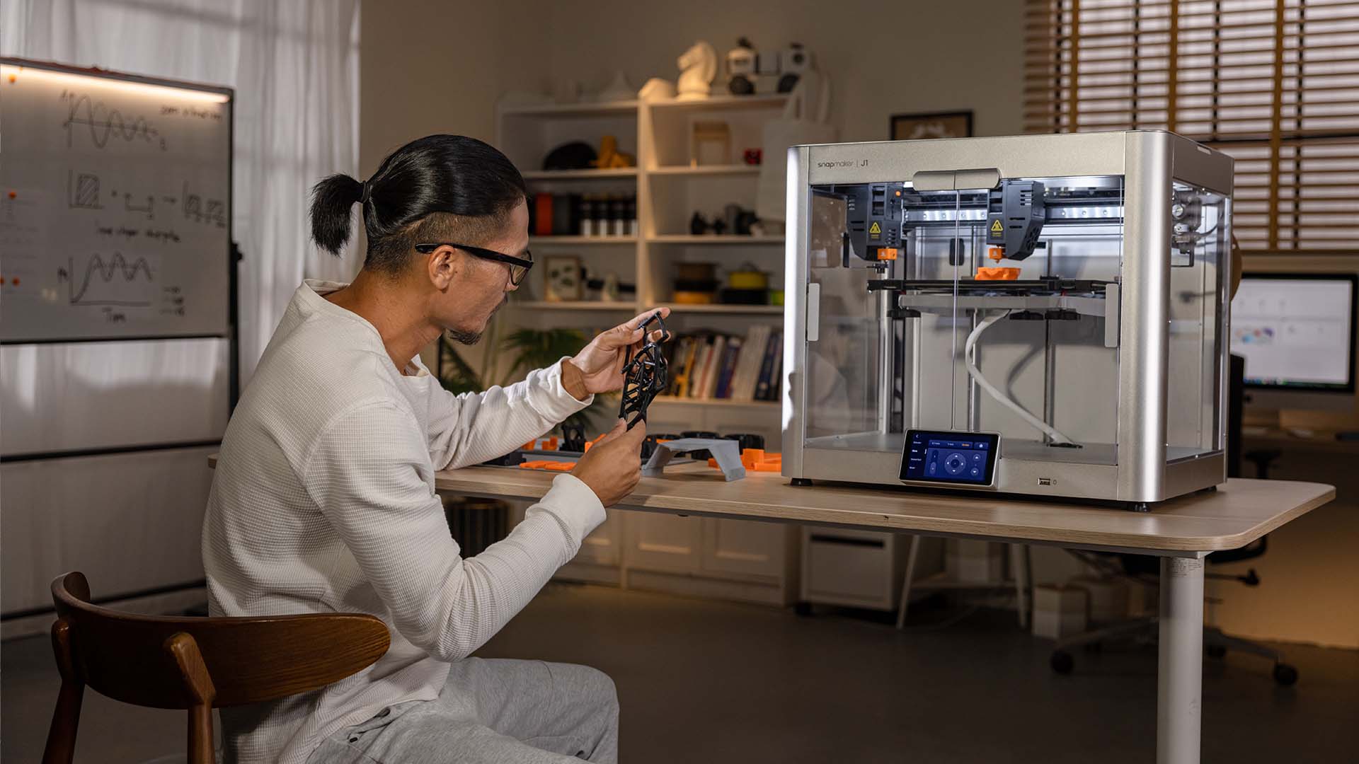 A person examines a printed object while seated next to a Snapmaker J1s 3D printer.