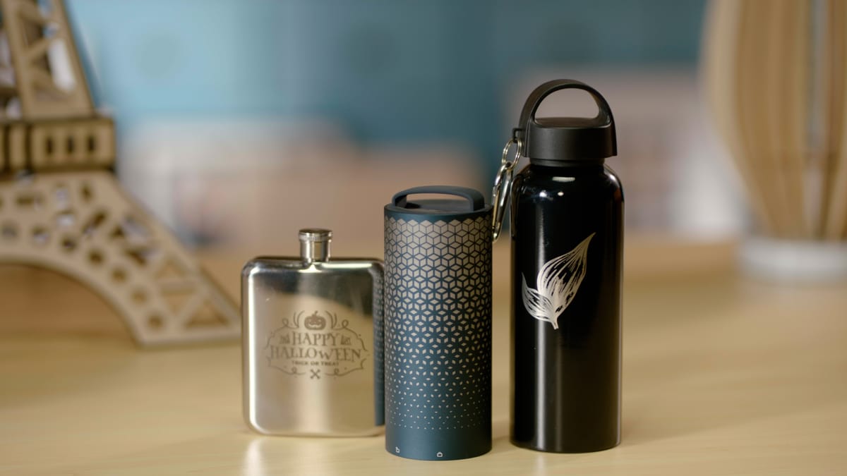Three laser-engraved tumblers displayed on a table, including a stainless steel flask with a Halloween design, a patterned blue tumbler, and a black bottle with a feather engraving.