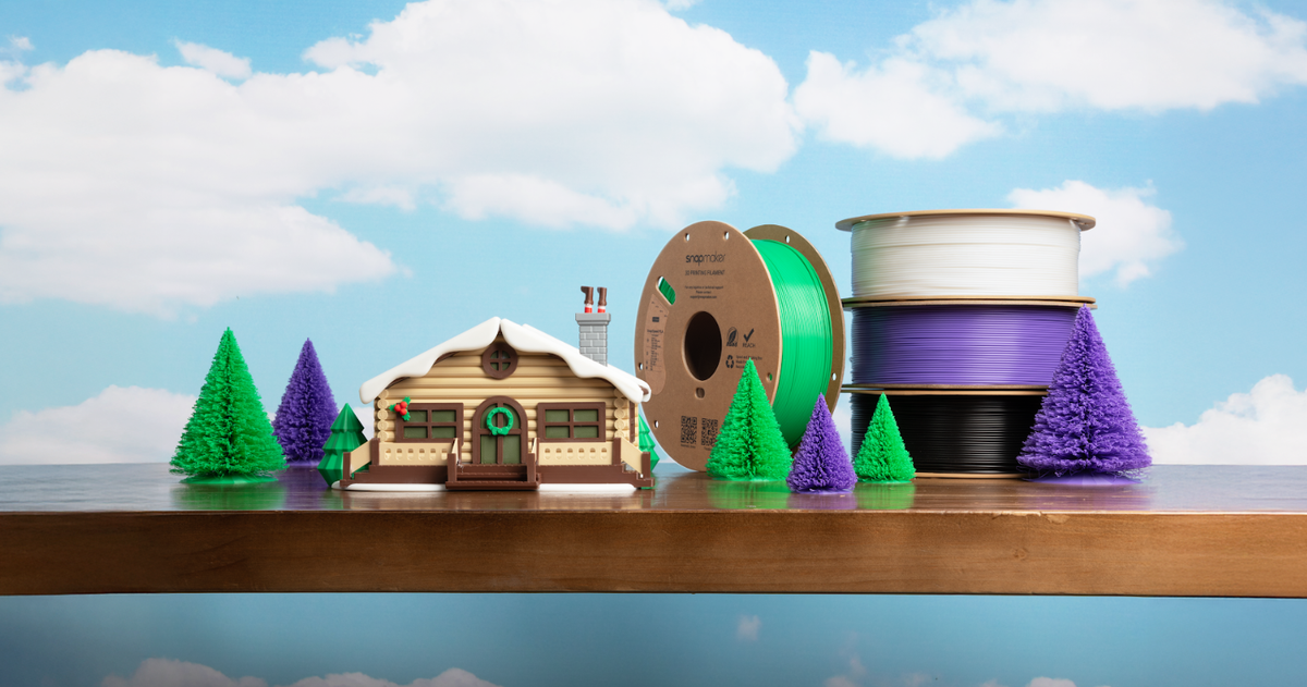 A small, detailed 3D printed model of a log cabin with miniature trees, arranged as a diorama on a wooden surface with a backdrop of blue sky and clouds.