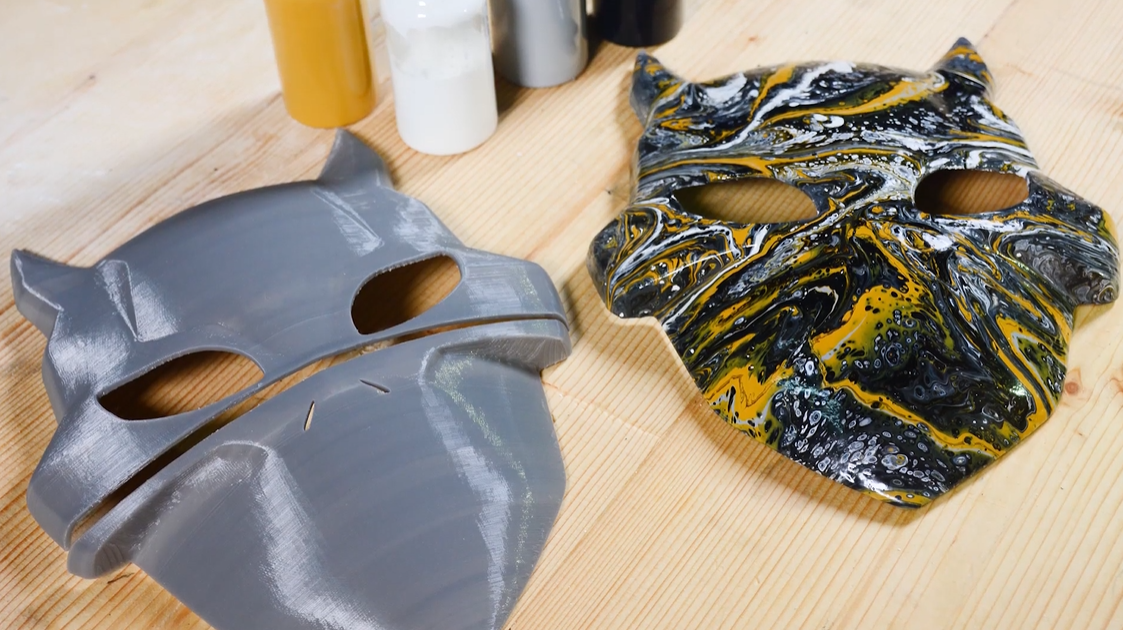 Two 3D printed masks are displayed side-by-side, one grey and unpainted, and the other vibrantly painted with black, white, and yellow swirls, illustrating the transformation.