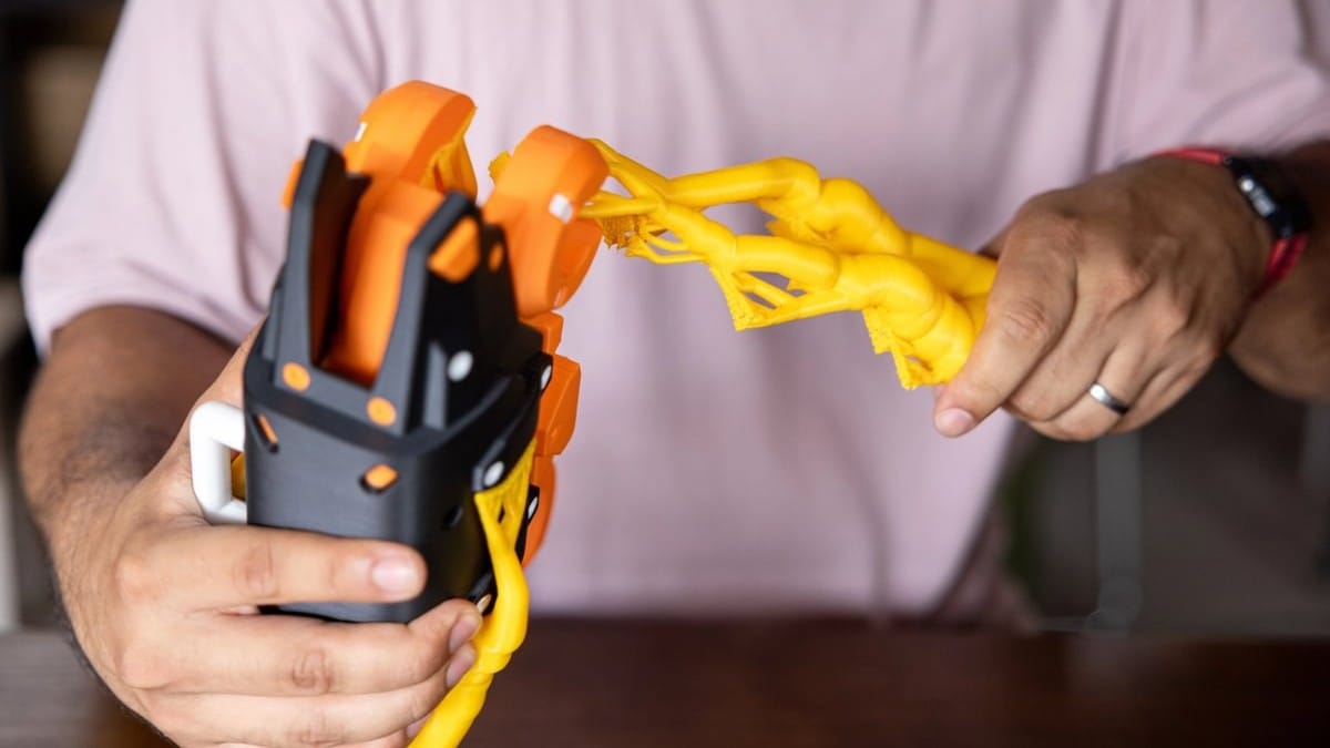 A person's hands carefully separating a bright yellow 3D printed support structure from a black and orange mechanical part.