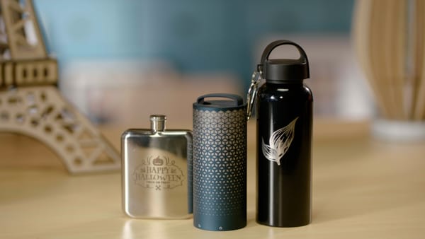Three laser-engraved tumblers displayed on a table, including a stainless steel flask with a Halloween design, a patterned blue tumbler, and a black bottle with a feather engraving.