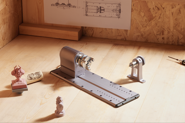 A CNC rotary module on a workbench, showcased with intricate carvings it produced, including a warrior bust and a lion statuette.