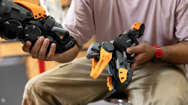 A person holding two complex, assembled 3D-printed robotic limbs made of black, grey, and orange plastic, showcasing the real-world strength and functional capabilities of FDM printed parts.